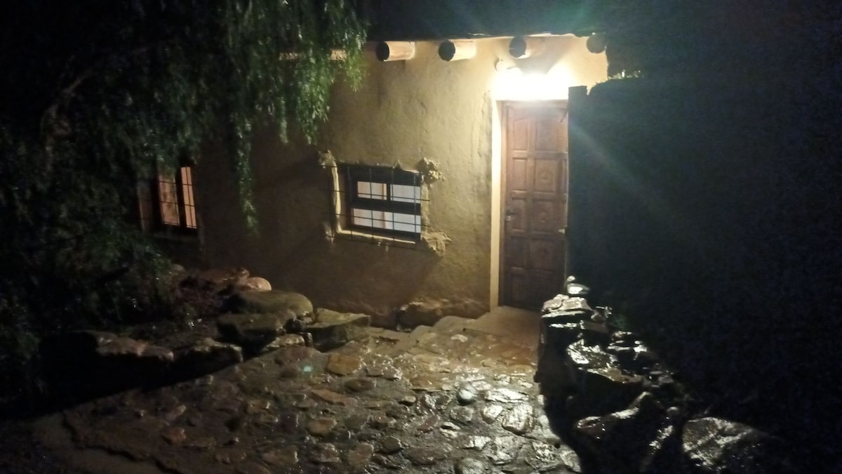 The entrance of a rustic dwelling is illuminated by a warm light, highlighting the textured stone pathway. A wooden door with intricate carvings stands prominently, framed by a small window featuring grid patterns. Lush greenery surrounds the entrance, creating a serene ambiance.