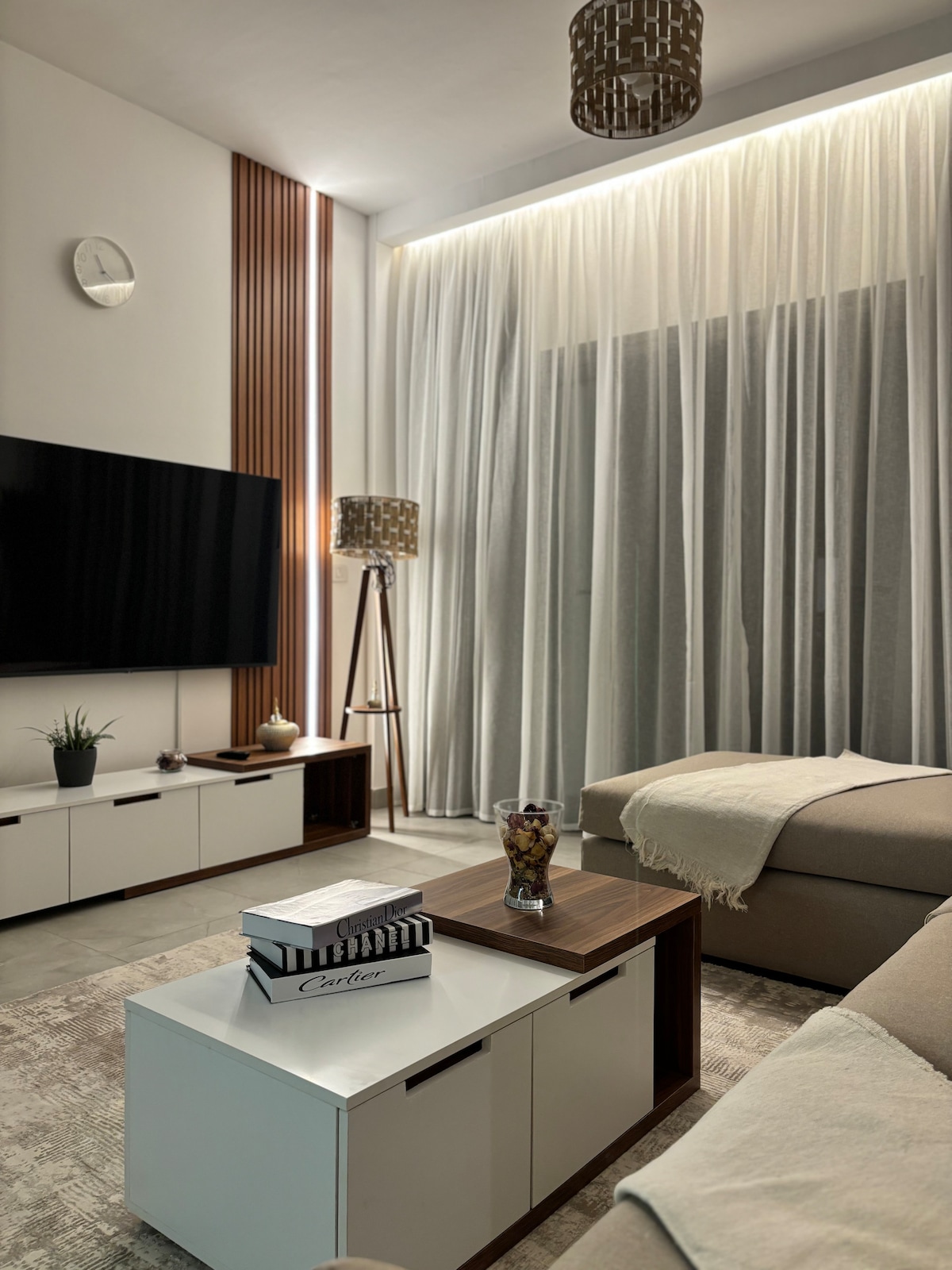A modern living area features a large flat-screen TV mounted on the wall, complemented by stylish decor. Natural light filters through sheer curtains, enhancing the tranquil ambiance. A plush sofa is positioned beside a wooden coffee table, topped with a decorative bowl and a book.