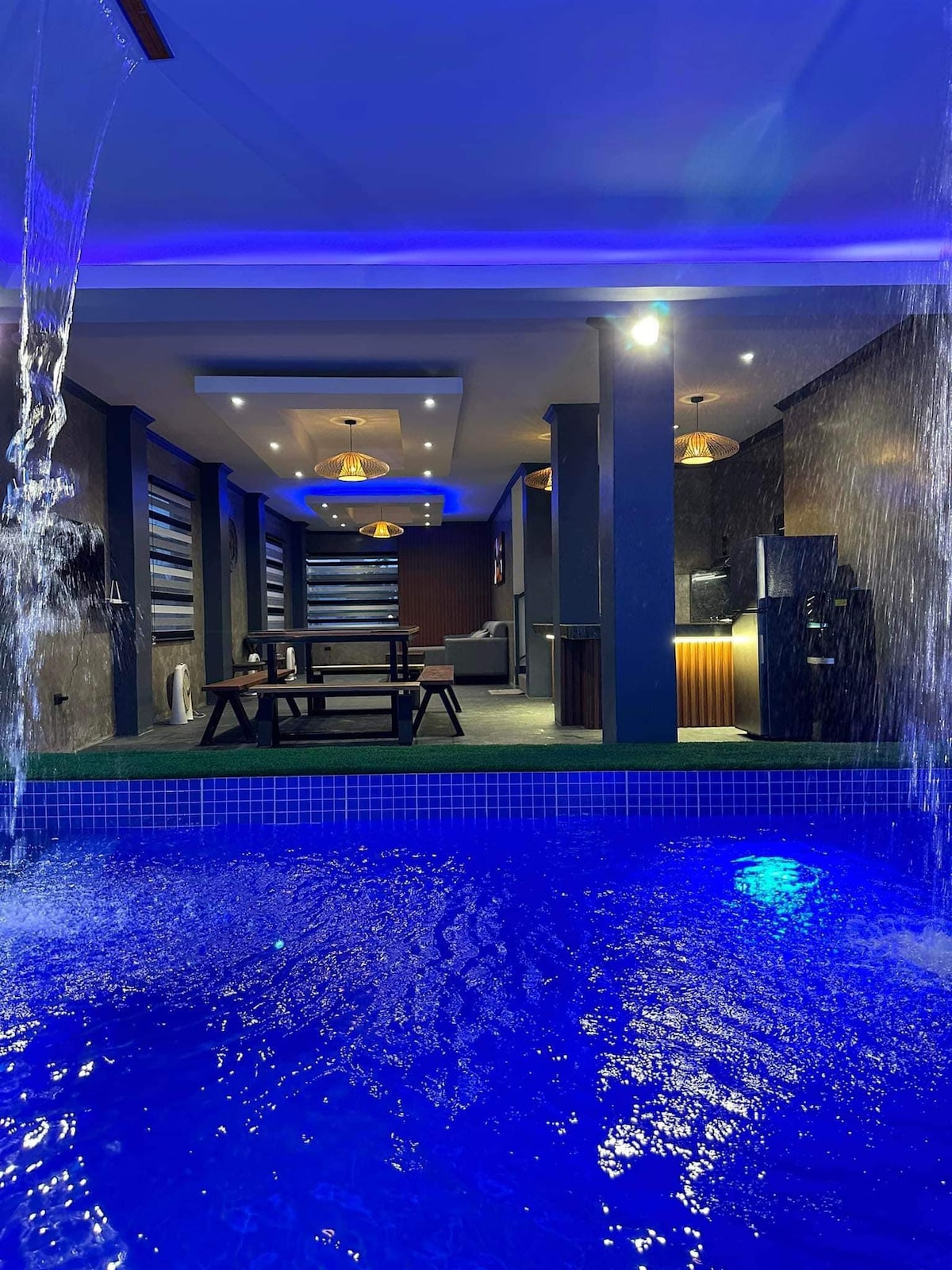 A spacious indoor area is highlighted by a shimmering blue pool, with water cascading from a feature wall. Modern pendant lights illuminate the space, showcasing comfortable seating and a dining area, while sleek kitchen amenities are visible in the background.