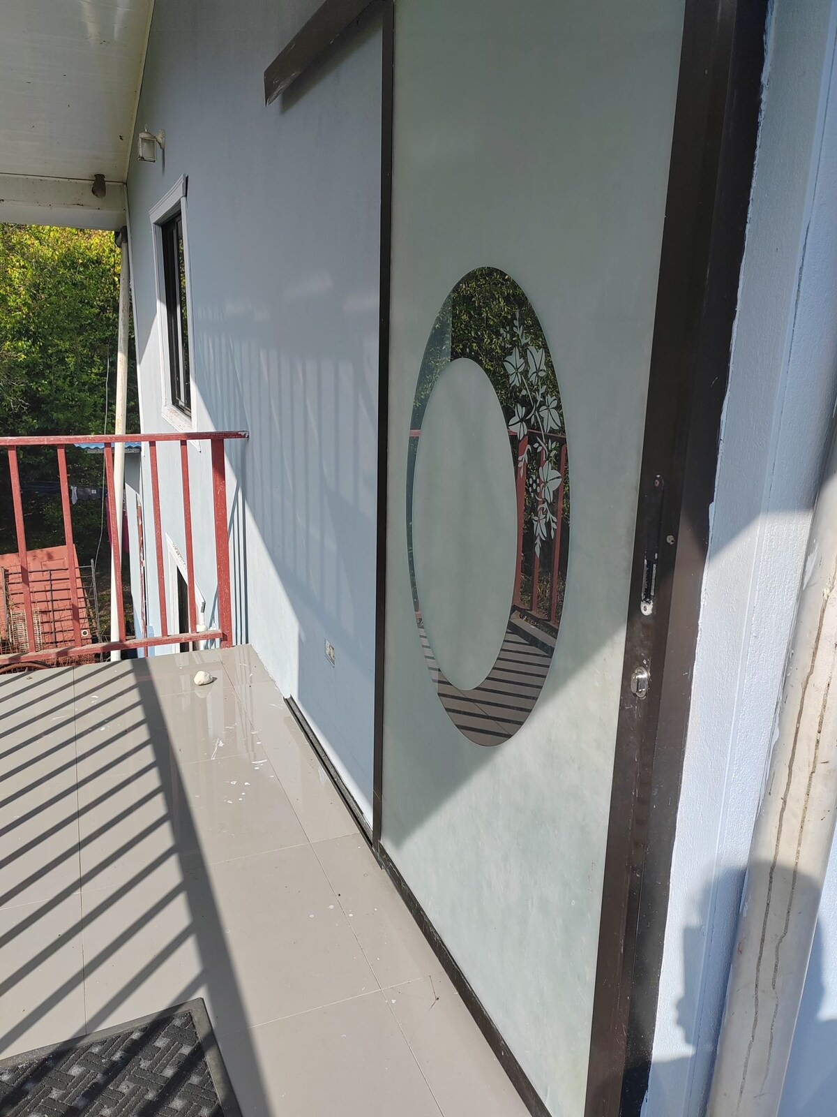 A balcony space is visible, featuring a large mirror with a circular design mounted on the wall. Sunlight casts shadows from the railing, highlighting clean lines. The floor is tiled and the wall is painted in soft hues, creating a calm and inviting entryway.