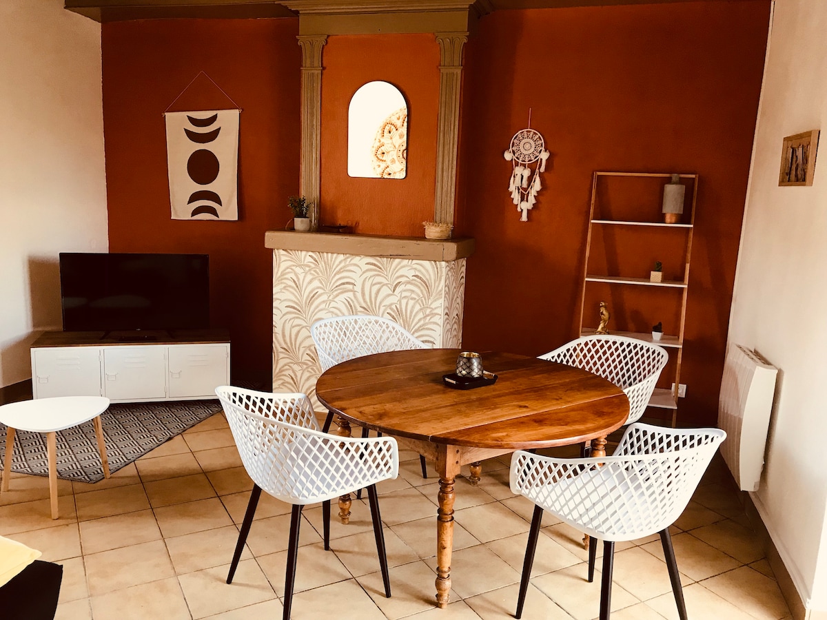 A dining area is presented with a round wooden table surrounded by four white chairs. A flat-screen television is positioned on a low storage unit nearby. Textured wall decor adds interest to the warm-colored walls, complemented by a decorative ladder shelf.