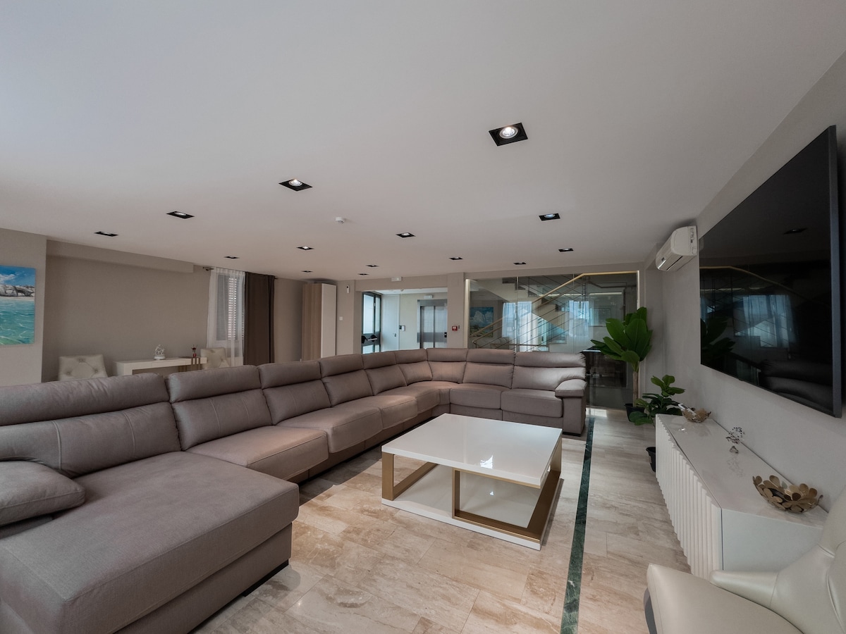 A spacious living area features a large sectional sofa in a neutral tone, paired with a modern coffee table at its center. Soft lighting is provided by recessed ceiling fixtures, and large windows allow natural light to fill the room. A decorative plant and TV are visible.