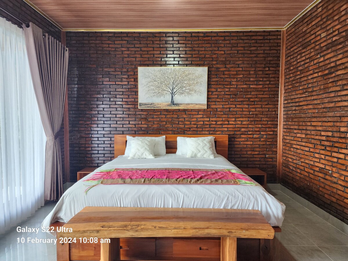 A cozy sleeping area features a spacious bed with white linens and a colorful blanket draped across the top. The brick walls provide a natural texture, while a large artwork of a tree is mounted above the bed. Light enters through sheer curtains beside the window.
