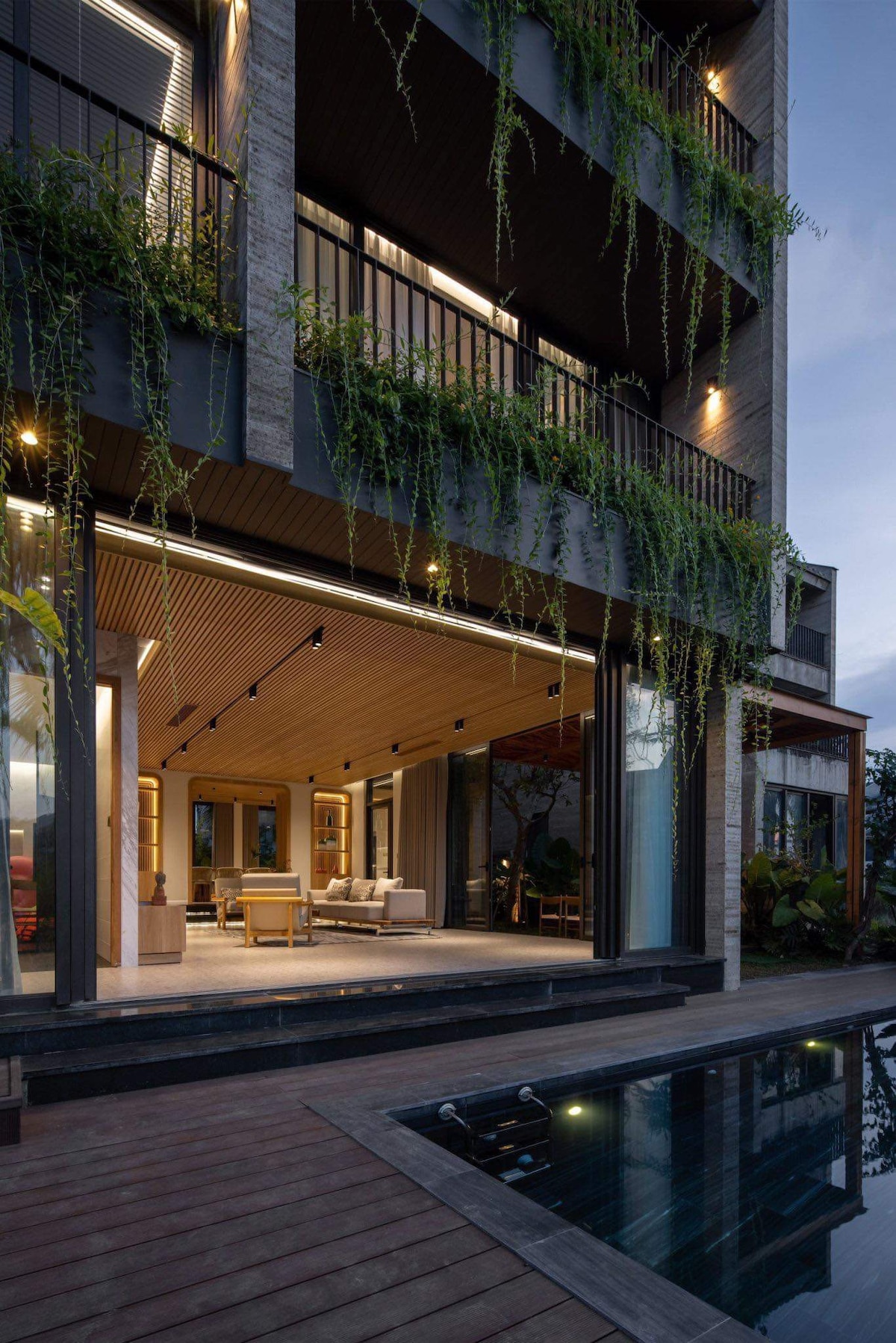A modern building exterior features large glass doors that open to a spacious patio. Soft lighting casts a warm glow on the furniture, while lush greenery hangs from the balcony. A tranquil pool is visible beside the wooden deck, enhancing the serene ambiance.