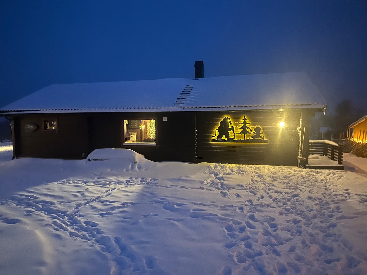 A charming wooden cabin is set against a snowy landscape under evening light. The exterior features decorative silhouettes of trees and figures, while soft illumination highlights the entrance. Snow blankets the ground, creating a serene atmosphere in the tranquil surroundings.