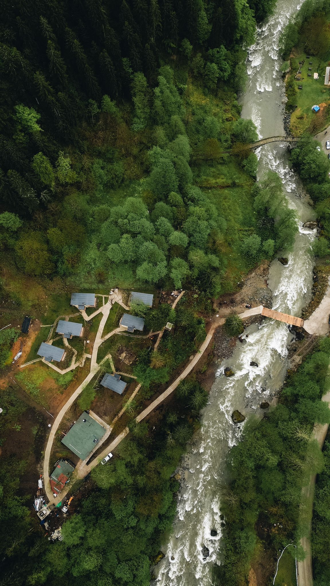An aerial view captures a serene landscape, featuring lush green trees surrounding several cottages along the riverbank. A winding path connects the structures, leading to a wooden bridge crossing the flowing river, reflecting the beauty of the natural environment.