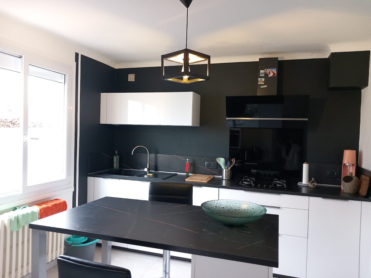 A modern kitchen is presented with a sleek black countertop and white cabinetry. Natural light filters through large windows, highlighting a round glass bowl on the dining table. Kitchen appliances and utensils are organized on the counter, contributing to a functional cooking space.