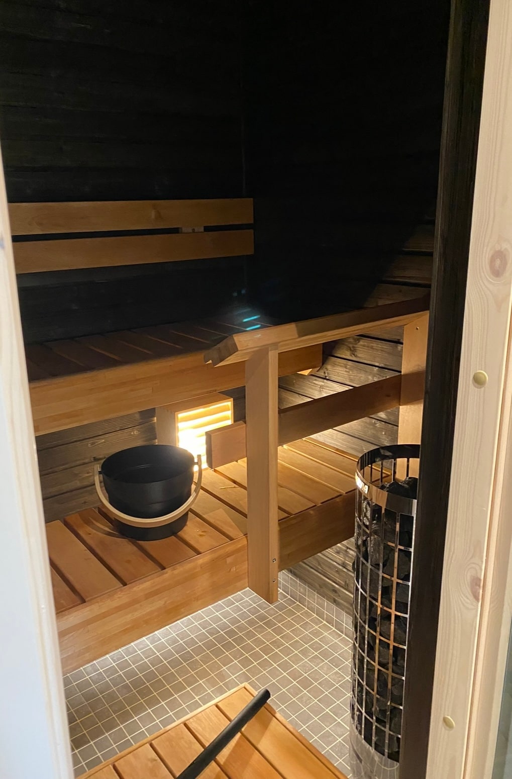 A wooden sauna interior is presented, featuring bench seating on multiple levels. A black bucket is positioned at the edge, and a metal stove stands beside it. Soft lighting enhances the warm wood tones, creating a calming atmosphere within the enclosed space.