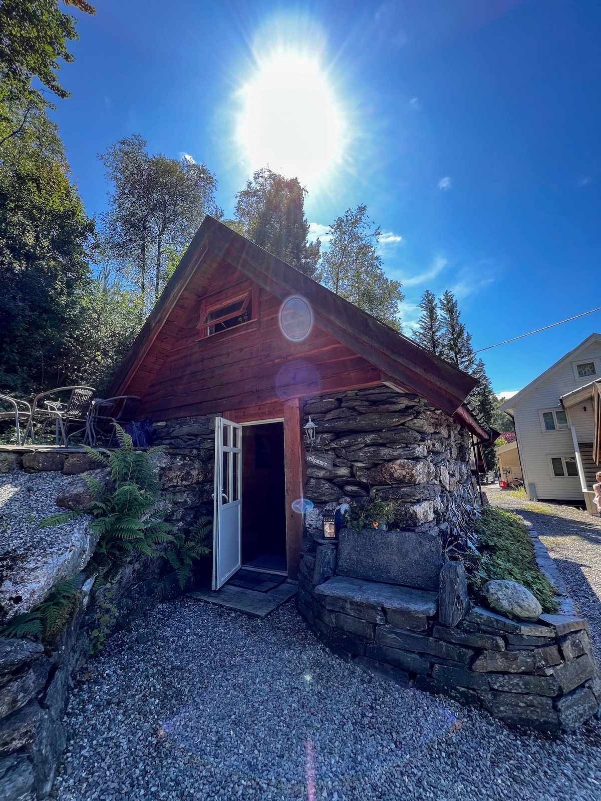 A charming cabin features a traditional wooden structure with a distinct sloped roof. Sunlight shines brightly from above, highlighting the surrounding greenery. A pathway leads to an open door, inviting guests to explore the interior. Natural stone accents provide a rustic feel.