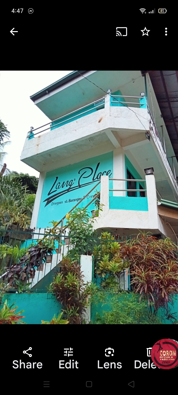 The exterior of the building features a two-story structure painted in blue and white. A welcoming sign is displayed on the wall, and lush greenery surrounds the entrance. A staircase leads to the upper level, framed by colorful plants and foliage.