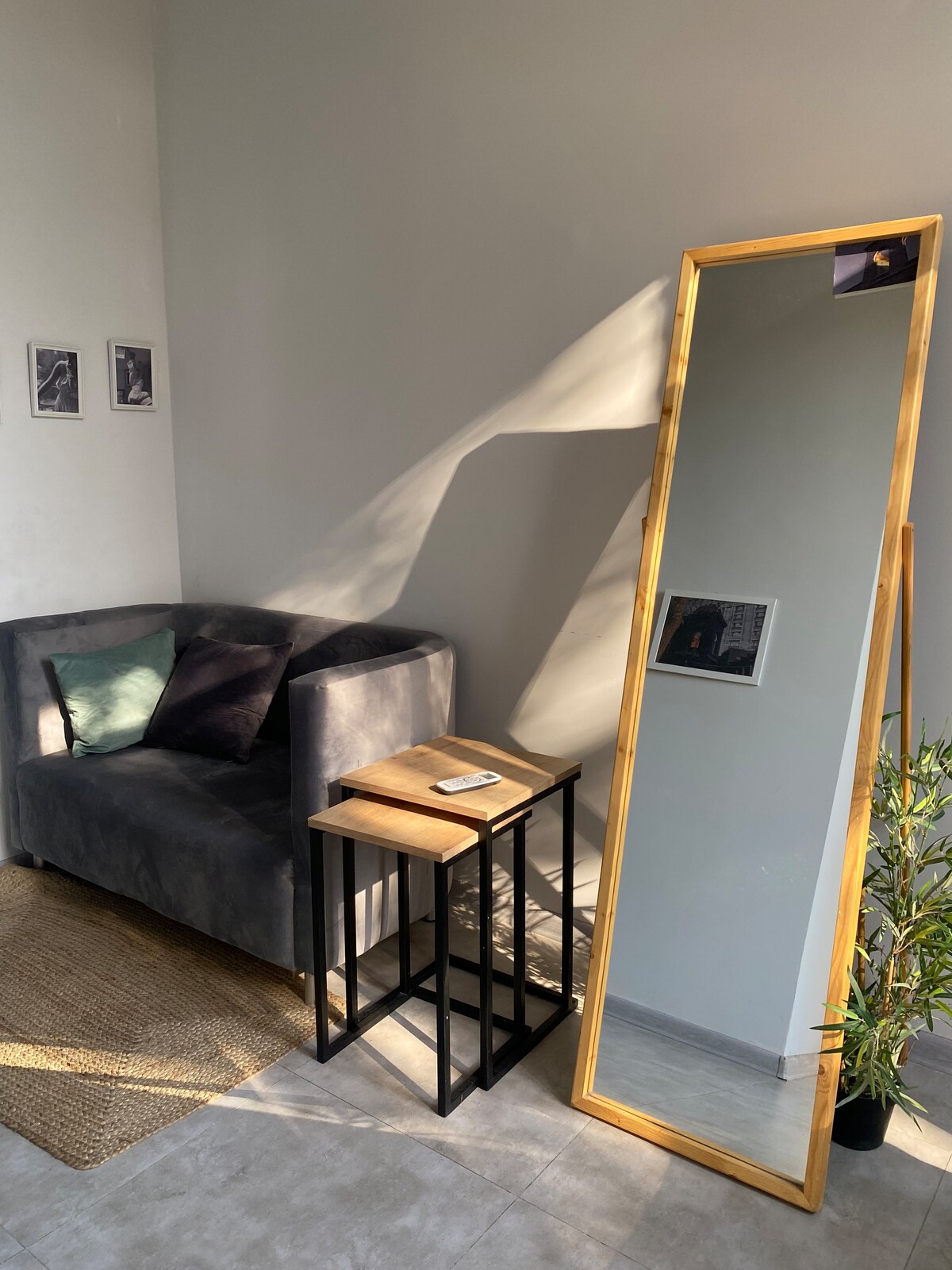 A cozy corner features a tall, standing mirror framed in wood, reflecting natural light. A dark gray sofa is paired with two small, wooden side tables. Decorative plants add a touch of greenery to the space, enhancing its inviting atmosphere.