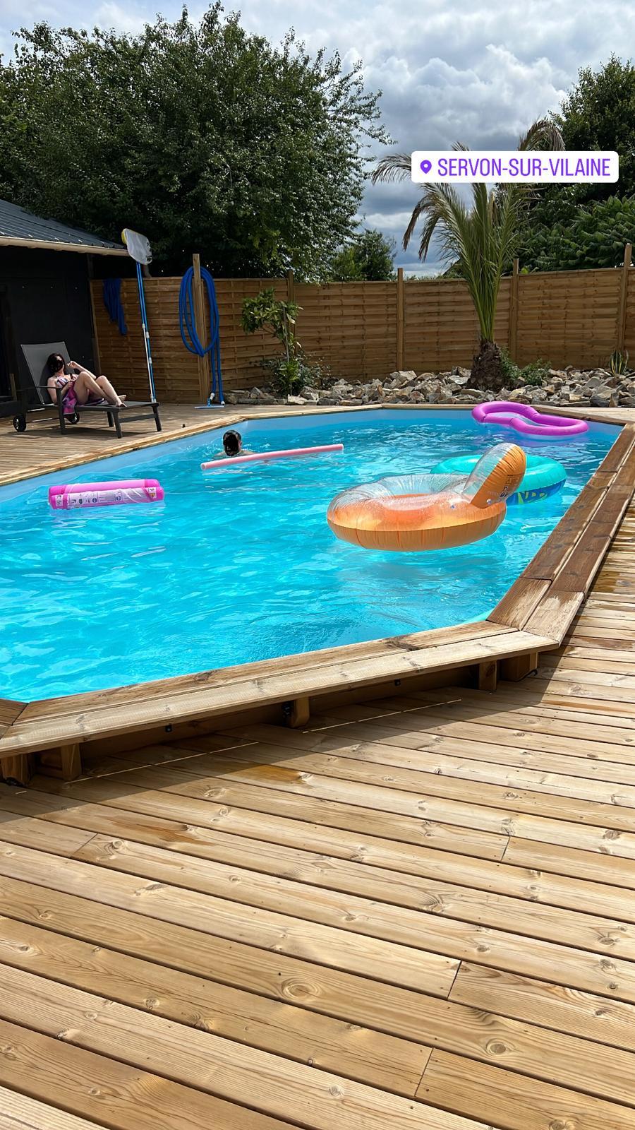 An inviting outdoor pool is surrounded by wooden decking and lush greenery. Several floating inflatables rest on the water's surface, while a single lounge chair is positioned nearby for relaxation. Soft clouds drift across the sky, enhancing the serene atmosphere.