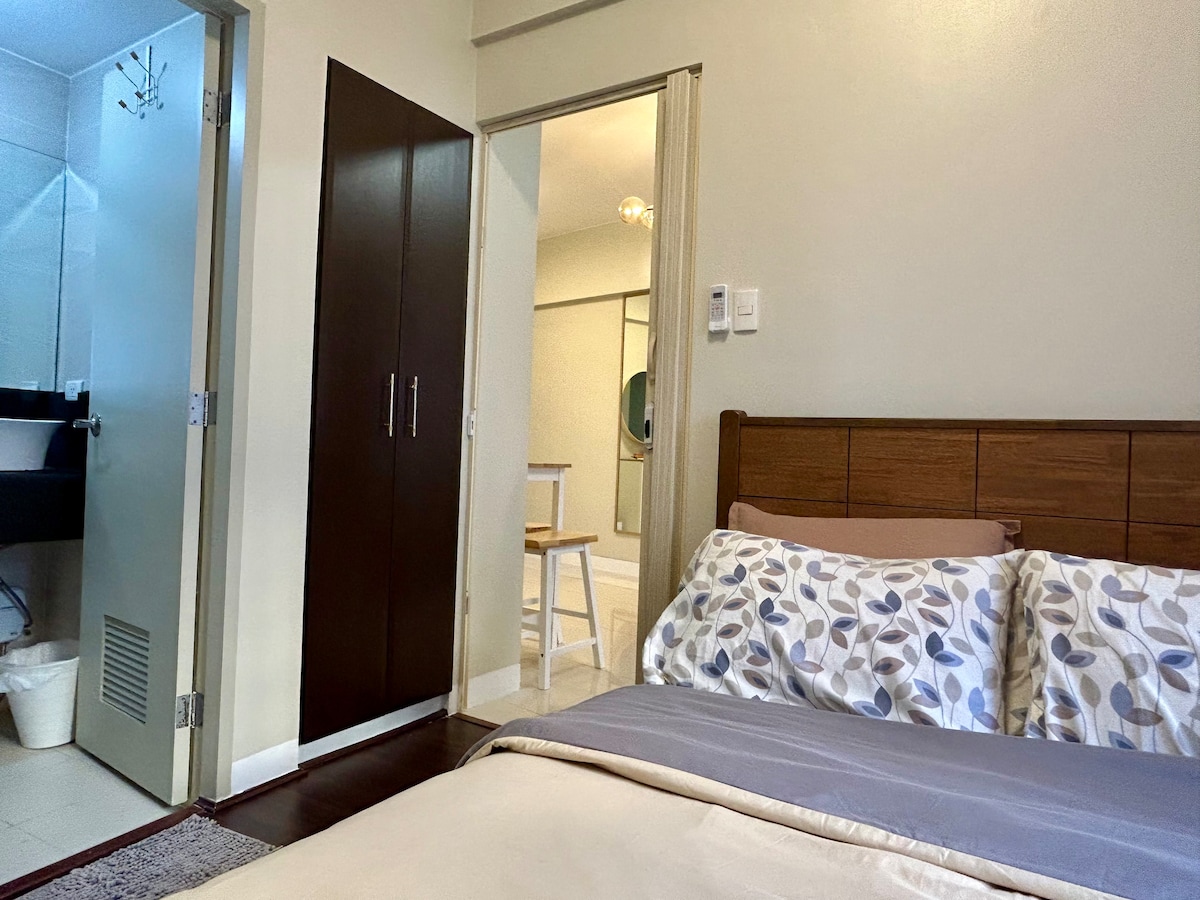 A queen bed adorned with patterned pillows is positioned against a light-colored wall. The room features doorways leading to a bathroom and closet space. Soft, neutral tones create a calming atmosphere, with natural light illuminating the area.