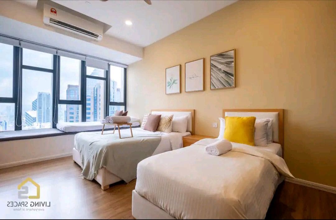A well-lit bedroom features two beds, one adorned with a white comforter and the other with a light green blanket. Large windows offer views of the city skyline, while three framed art pieces enhance the wall above the beds.