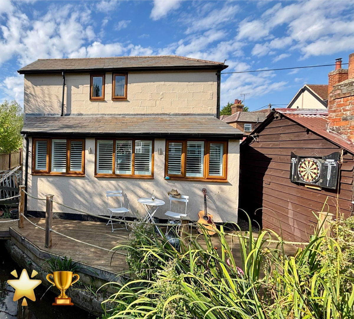 The exterior of Little Brook Retreat features a charming two-story building with large windows and a deck area. Small white tables and chairs are visible on the deck, accompanied by greenery. A wooden dartboard is mounted on a nearby shed, enhancing the outdoor space.