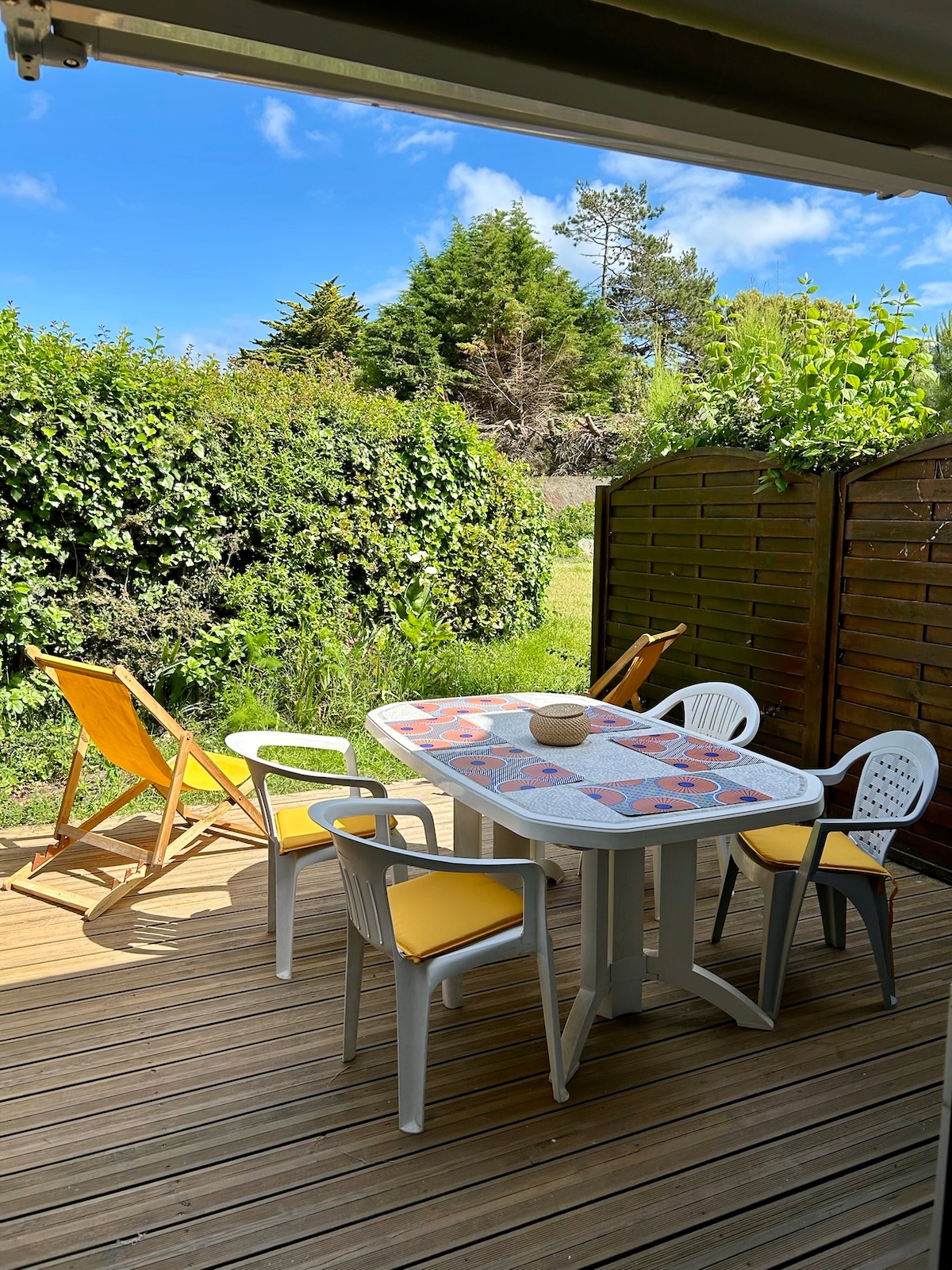 A private terrace features a wooden deck adorned with a dining table and six chairs. Two loungers sit nearby, surrounded by lush green foliage. The area is illuminated by natural light, creating a welcoming outdoor space for meals or relaxation.
