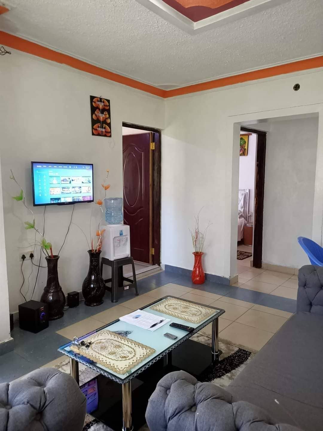 A cozy living area is depicted, featuring a comfortable sofa and a glass coffee table. A television is mounted on the wall, and a water cooler is situated nearby. Decorative vases with plants add a touch of color, enhancing the welcoming ambiance.