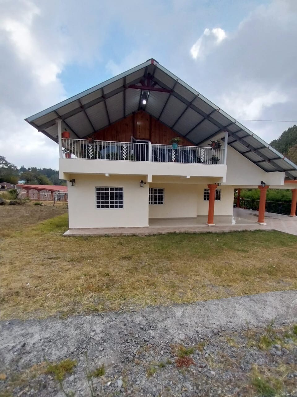 A two-story villa is showcased with a sloped roof, featuring a balcony adorned with plants. Large windows are present, providing natural light to the interior. The exterior is complemented by a grassy area and a stone pathway leading up to the entrance.
