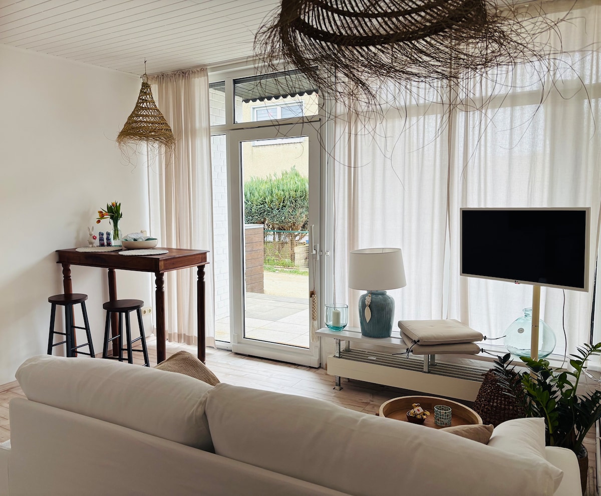 A bright living area is observed, featuring large windows with sheer curtains that allow natural light to filter in. A comfortable sofa faces a television on a modern console, while a small wooden bar with stools is positioned nearby, enhancing the inviting space.