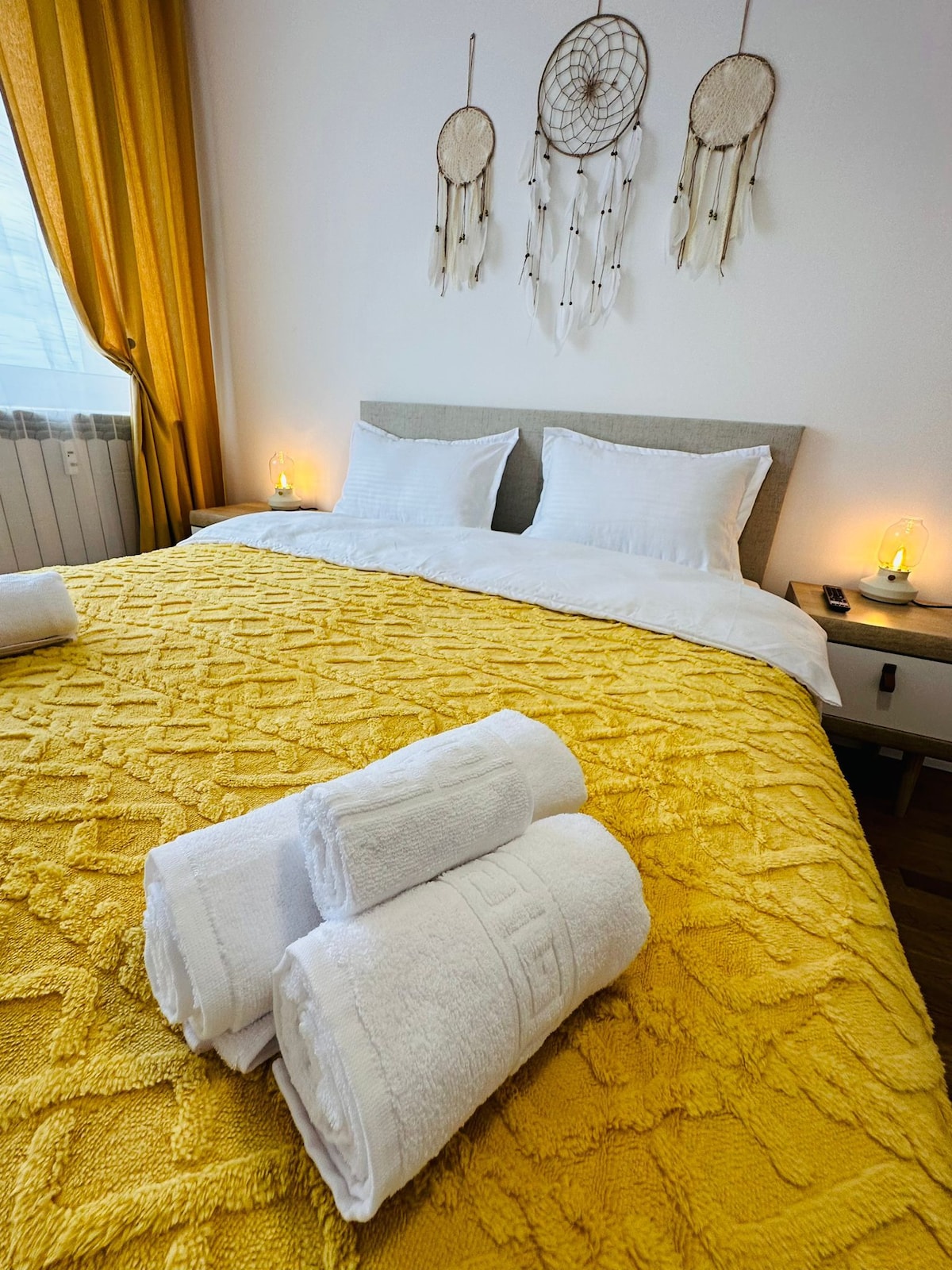 A spacious bed with a textured yellow blanket is complemented by a light grey headboard. White towels are neatly arranged at the front. Dreamcatchers adorn the wall above the bed, and natural light filters through sheer curtains, creating a soft ambiance.