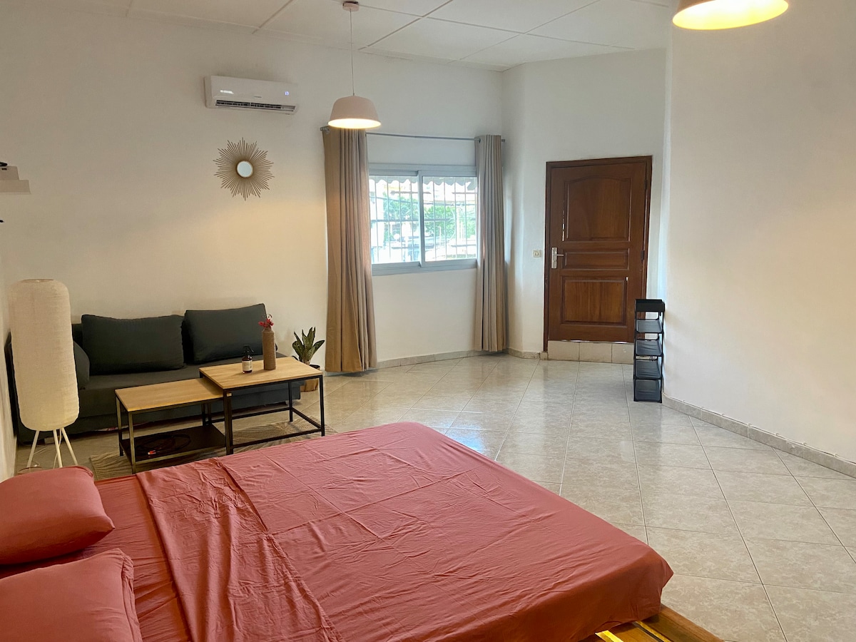 An open studio space features a spacious bed with a warm red coverlet. A green sofa sits across from the bed, accompanied by a small side table. Natural light filters through a window covered with light curtains, illuminating the room's neutral palette.