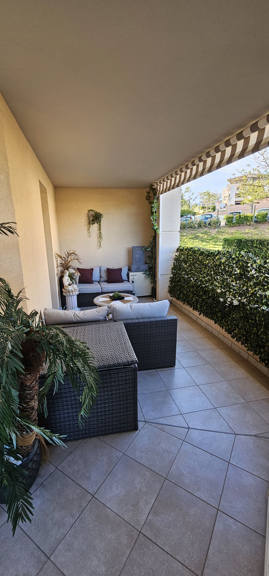 A covered terrace features two comfortable seating areas with cushions, complemented by potted greenery. The tiled floor reflects natural light, while a decorative privacy hedge adds a touch of serenity. The space is designed for relaxation in a peaceful environment.