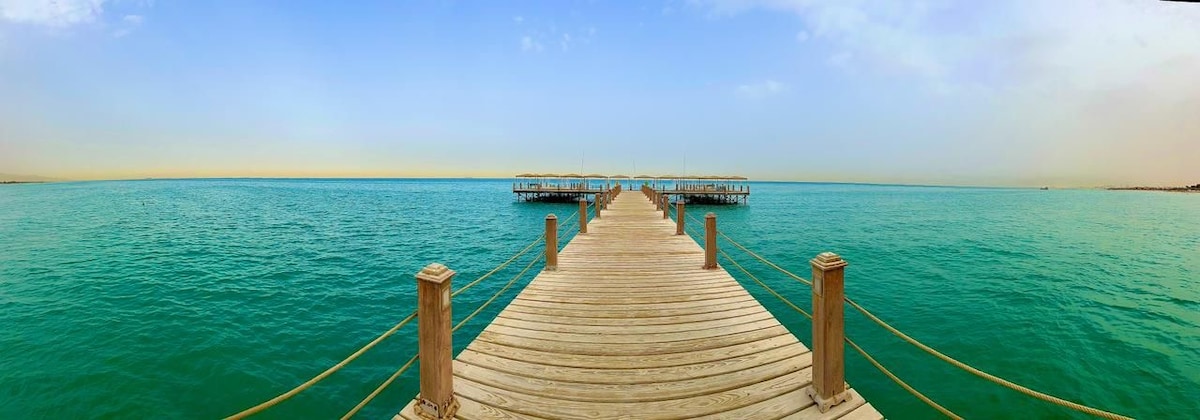 A wooden pier extends into a calm, turquoise sea, framed by a clear sky. The structure features railings along its sides and is lined with shaded areas at the end, providing a serene atmosphere for relaxation.