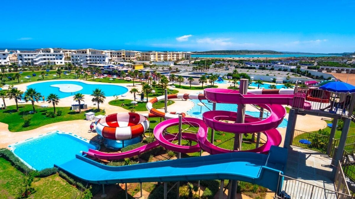 A wide view of a vibrant water park features colorful water slides and multiple swimming pools, surrounded by palm trees and well-kept greenery. A clear blue sky and distant coastline are visible, adding a refreshing backdrop to the leisure space.
