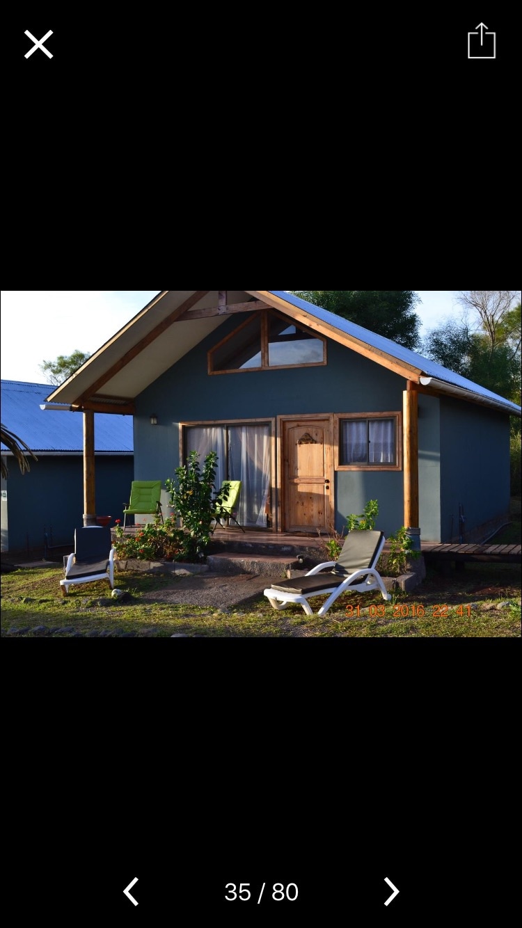 A charming cabin with a triangular roof is shown, featuring a wooden front door and large windows. Two green chairs and a lounging chair are positioned on the porch, surrounded by lush greenery and a well-maintained garden area.