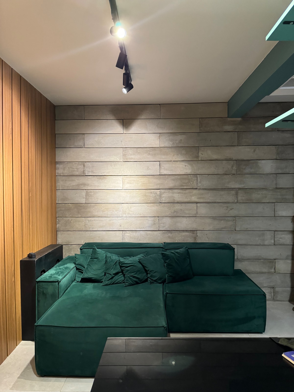 A section of a cozy living area features a green velvet sofa adorned with several matching cushions. The backdrop consists of a textured wall combining wooden panels and smooth stone, providing a modern, stylish ambiance. A dark coffee table is positioned in front.