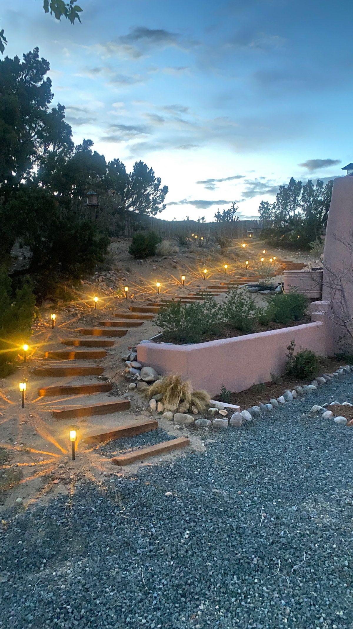 A softly lit pathway winds its way up a terraced area, bordered by native plants. Path lights illuminate the steps, creating a serene ambiance. The surrounding landscape features natural elements, including boulders and gravel, under a twilight sky.