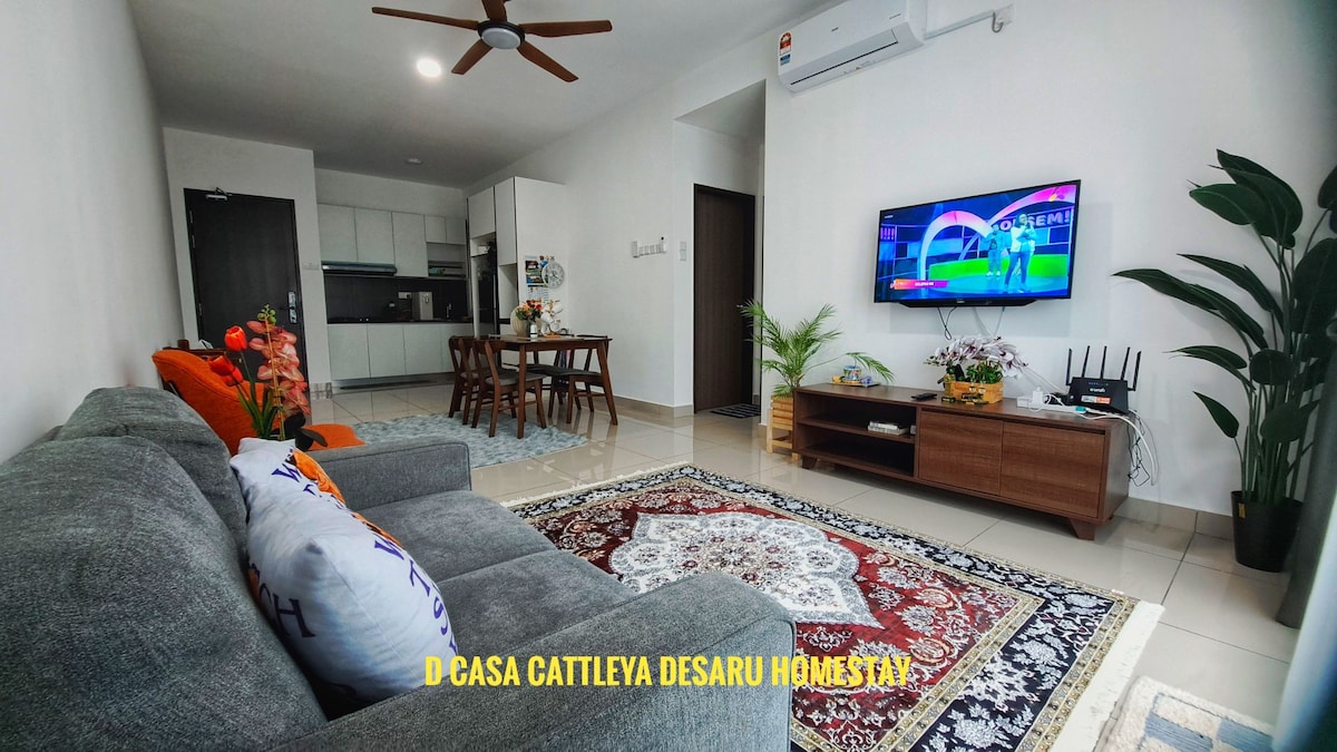 A spacious living area is presented with a comfortable gray sofa and plush throw pillows. A large television is mounted on the wall across from the seating. A wooden coffee table sits atop a decorative rug, while a dining area with a table and chairs is visible in the background.