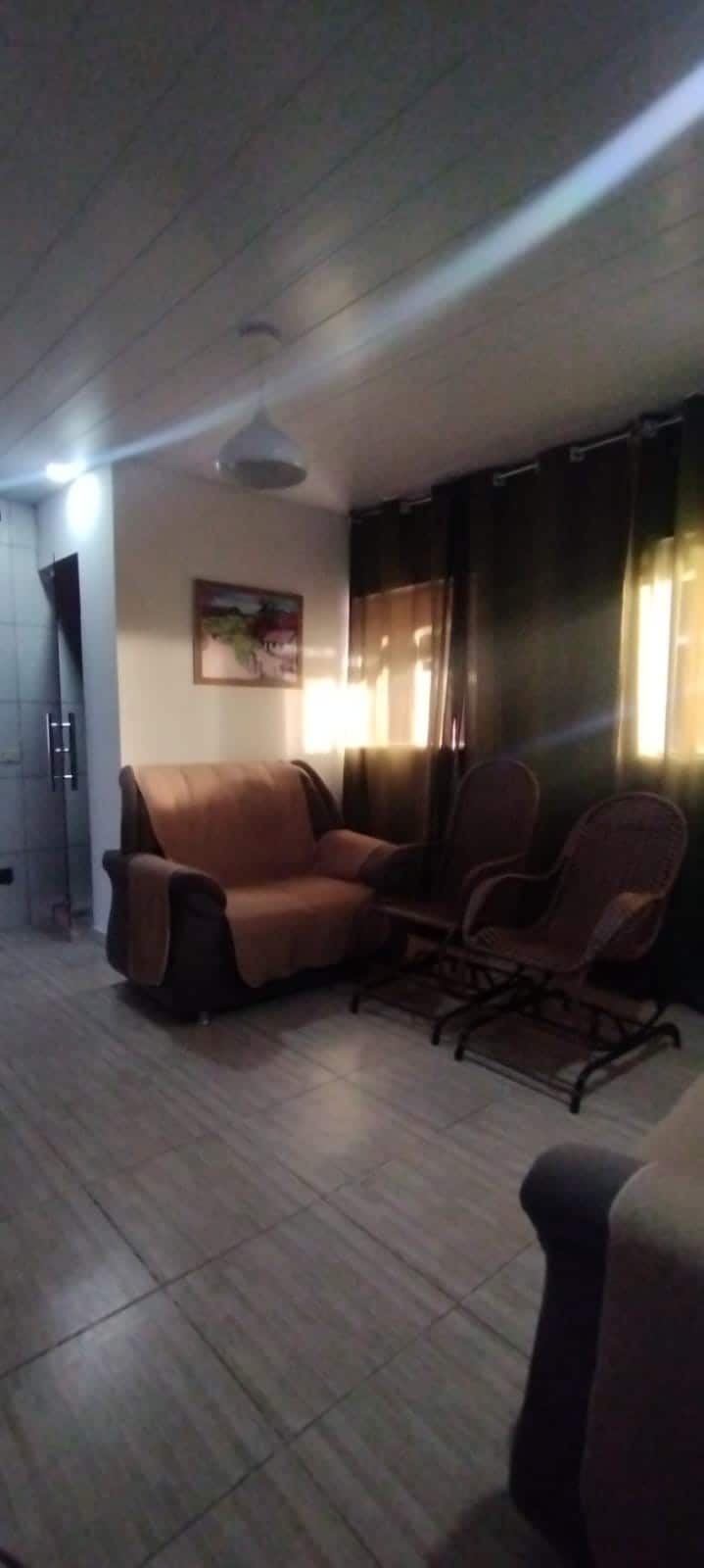A cozy seating area is presented with a brown couch and two rocking chairs positioned beside large windows. Natural light filters through sheer curtains, creating a warm and inviting space. A small piece of artwork is displayed on the wall, adding a touch of charm.