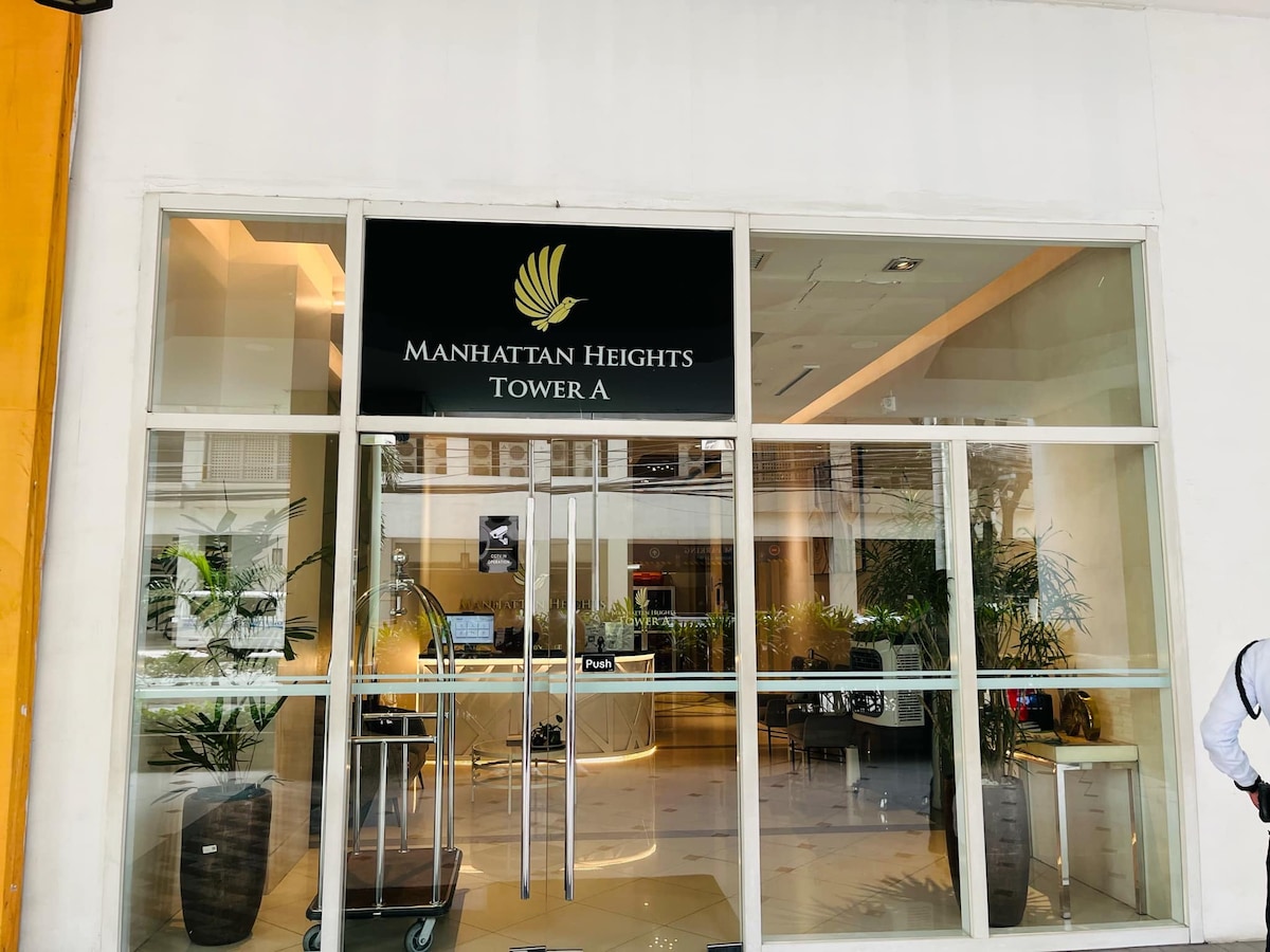 The entrance to Manhattan Heights Tower A is visible, featuring glass doors and a prominent sign. Potted plants are arranged on either side of the entrance, enhancing the modern aesthetic of the building's facade.
