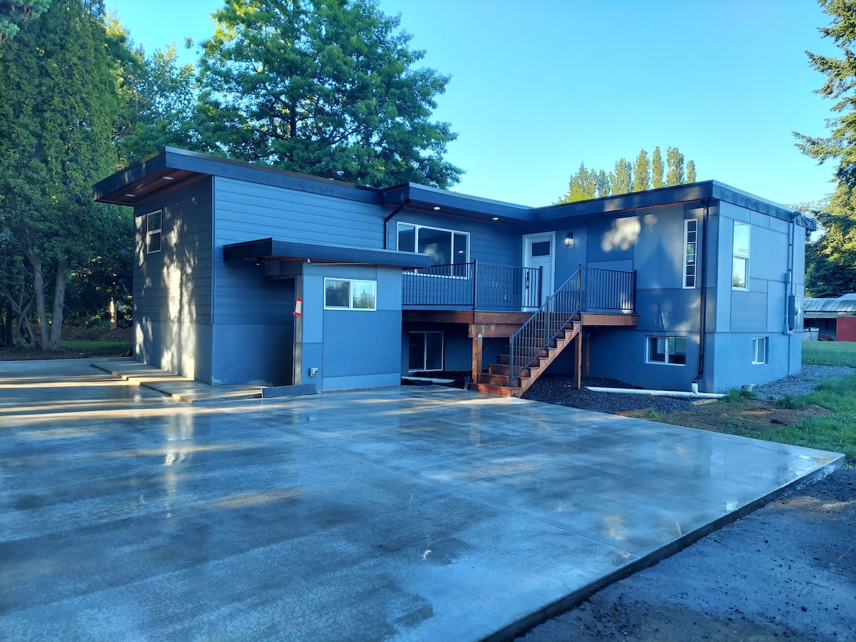 A modern two-story building exterior is shown, featuring a combination of light blue and dark gray siding. A wooden deck with steps leads to the entrance. A spacious concrete area is visible in front, surrounded by greenery and trees.
