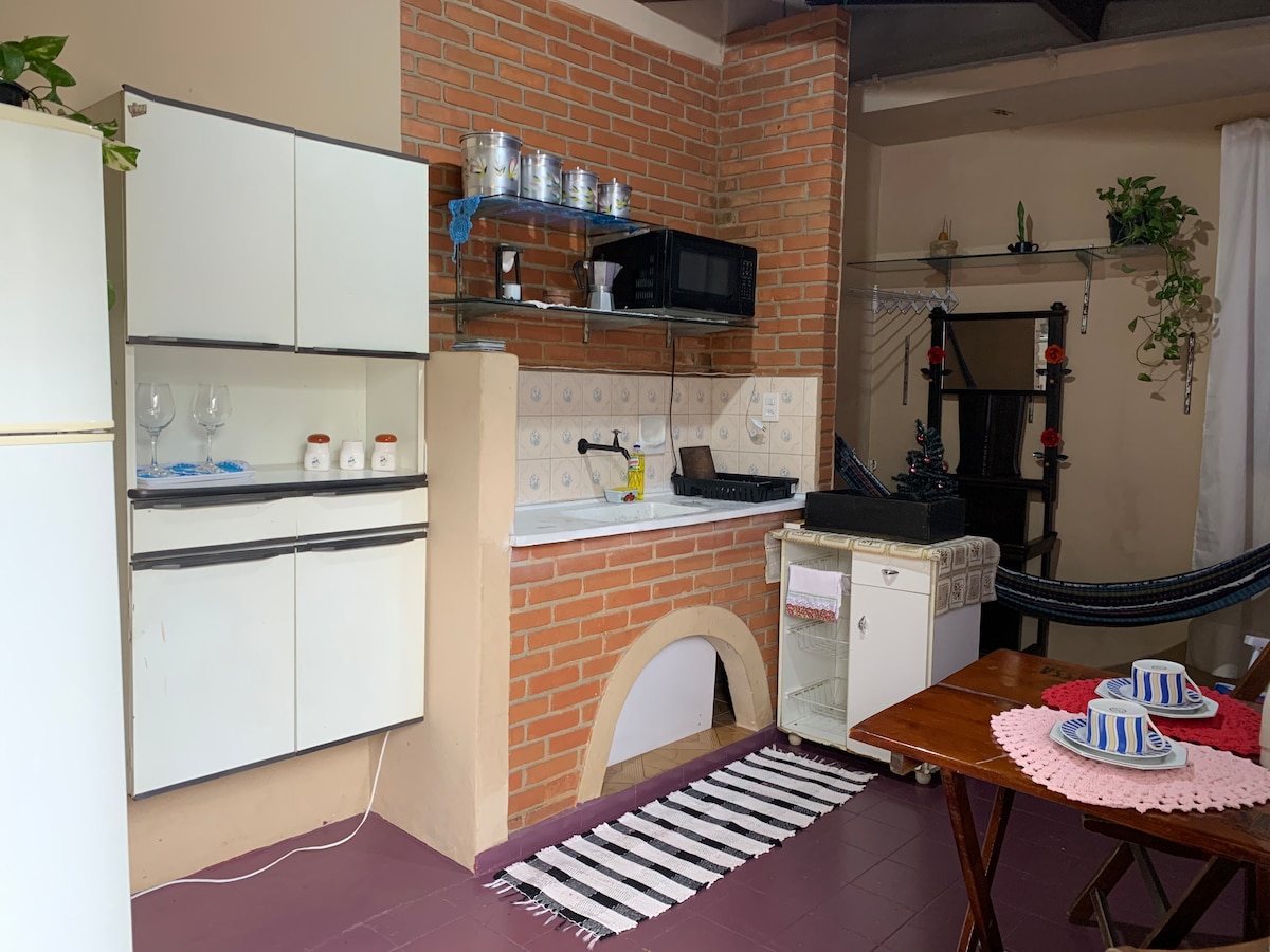 The kitchen area features a brick wall and white cabinets. A microwave and a sink are installed beneath the shelves holding glassware and utensils. In the foreground, a small dining table with two place settings and colorful coasters is accompanied by a white hammock, adding a relaxed feel to the space.
