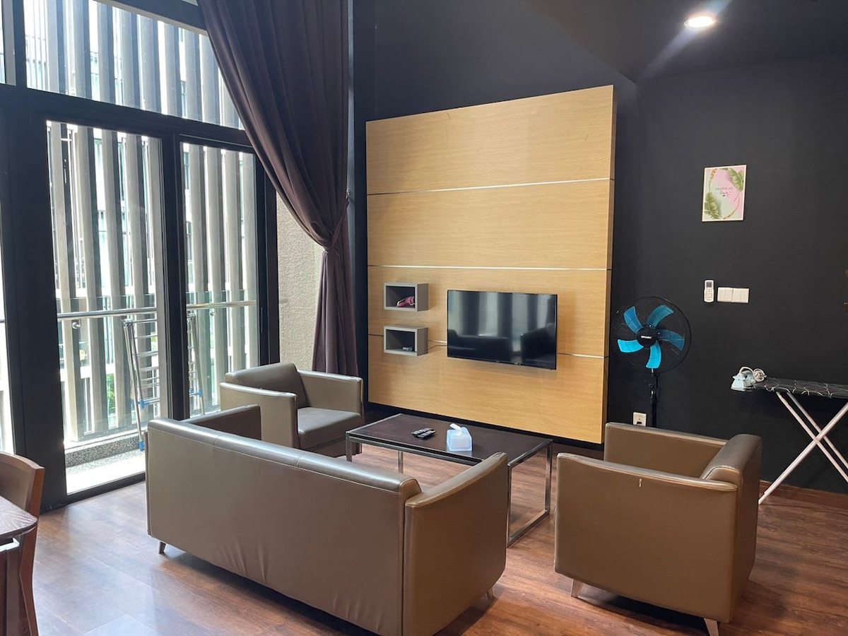 A modern living area features two brown leather sofas arranged around a central coffee table. A wall-mounted television is positioned above a sleek wooden panel. Natural light filters in through tall glass doors, complemented by dark curtains, while a ceiling fan provides comfort.