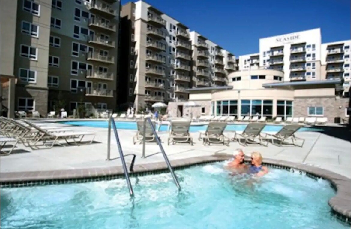 The image displays a resort area featuring a large pool surrounded by multiple lounge chairs. Two individuals are seen enjoying the hot tub, while the backdrop consists of a multi-story building with large windows, contributing to a vibrant resort atmosphere.