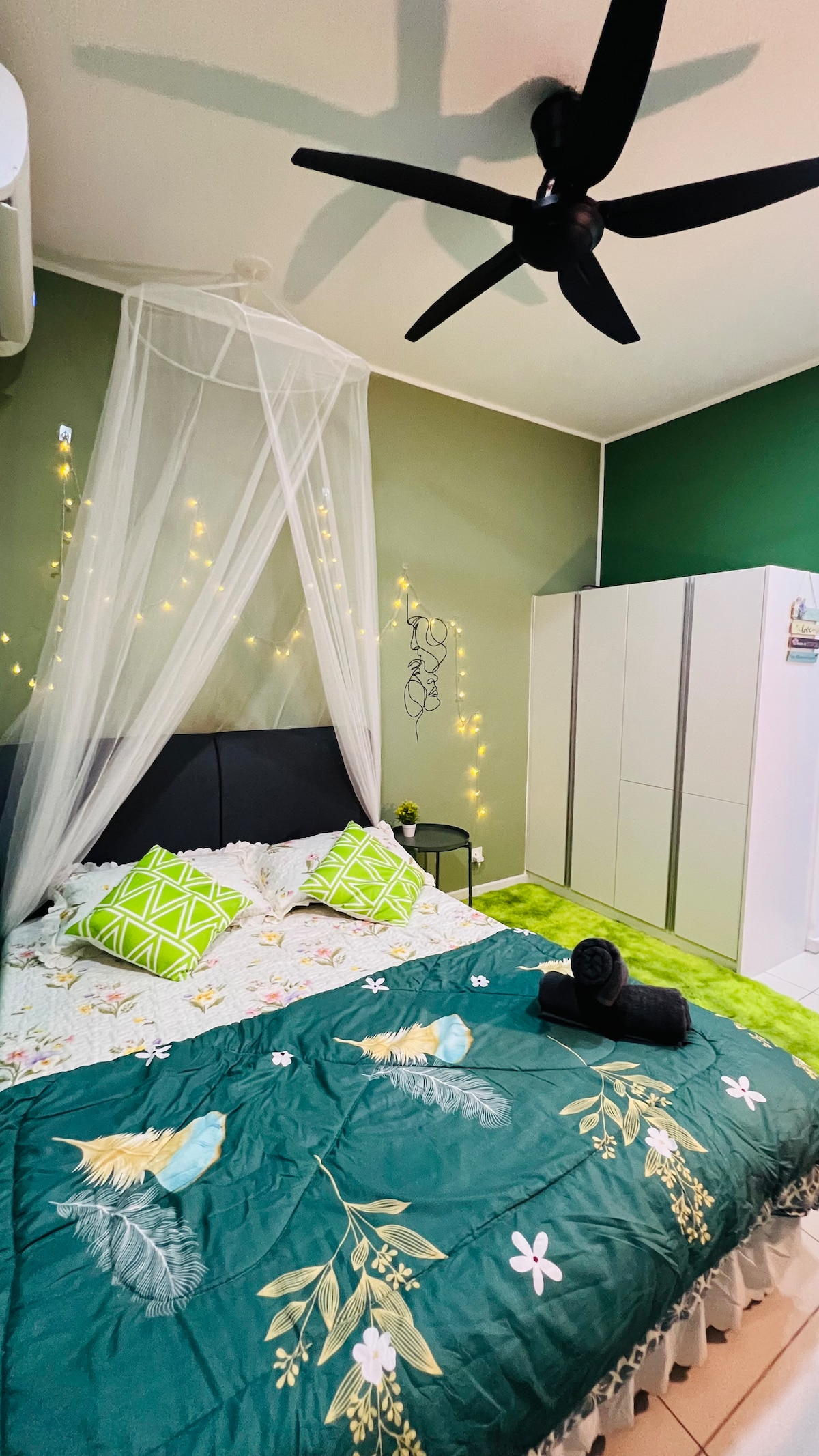 A cozy sleeping area features a spacious bed adorned with a vibrant green blanket and decorative pillows. Soft fairy lights are hung on the walls, adding a gentle glow. A wardrobe stands nearby, providing ample storage space.