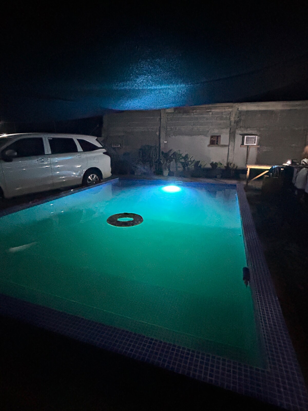 A calm night scene of a private pool is visible, featuring soft blue lighting that creates a serene atmosphere. The pool is bordered by a safety net overhead, with a vehicle parked nearby, and plants enhance the surroundings.