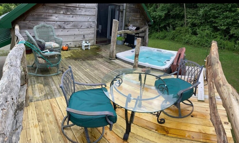 An outdoor deck features a hot tub, surrounded by wooden railings and lush greenery. A glass-top table is accompanied by two swivel chairs with green cushions. Two lounge chairs are positioned nearby, creating a comfortable space for relaxation.