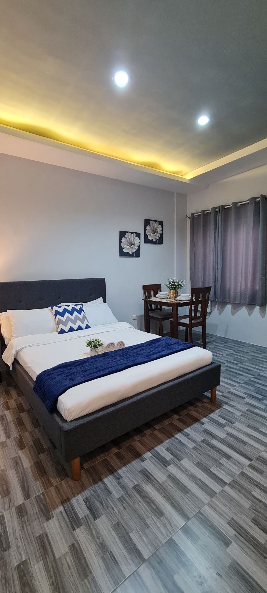 A cozy room features a spacious bed with a soft blue blanket and decorative pillows. A small dining table with two chairs is positioned near the window, which is draped with gray curtains. Wall art depicting flowers adds a touch of decor.