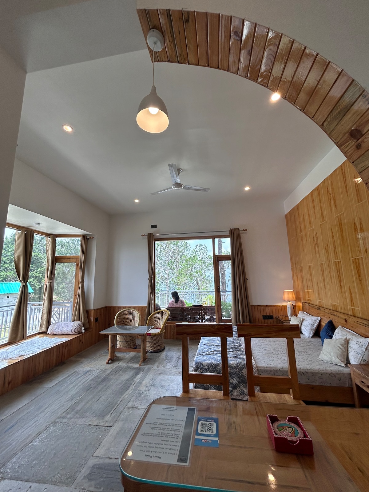 A bright room features large windows that offer views of the surrounding greenery. Two wicker chairs are positioned beside a wooden table. A cozy seating area is enhanced by warm wooden accents, while soft lighting is provided by a ceiling fixture, complementing the natural light.