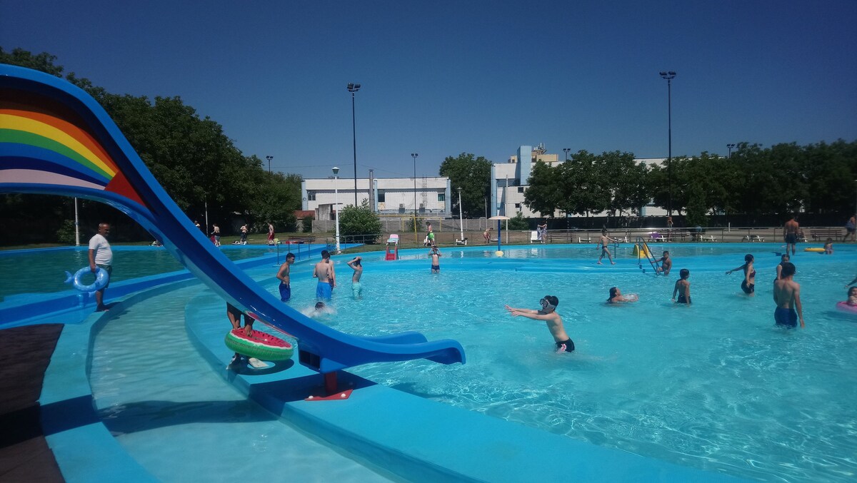 A vibrant pool area features children and adults enjoying the water on a sunny day. A colorful water slide with a rainbow design stands prominently, and pool floats are scattered throughout the sparkling blue water. Surrounding greenery adds to the refreshing atmosphere.