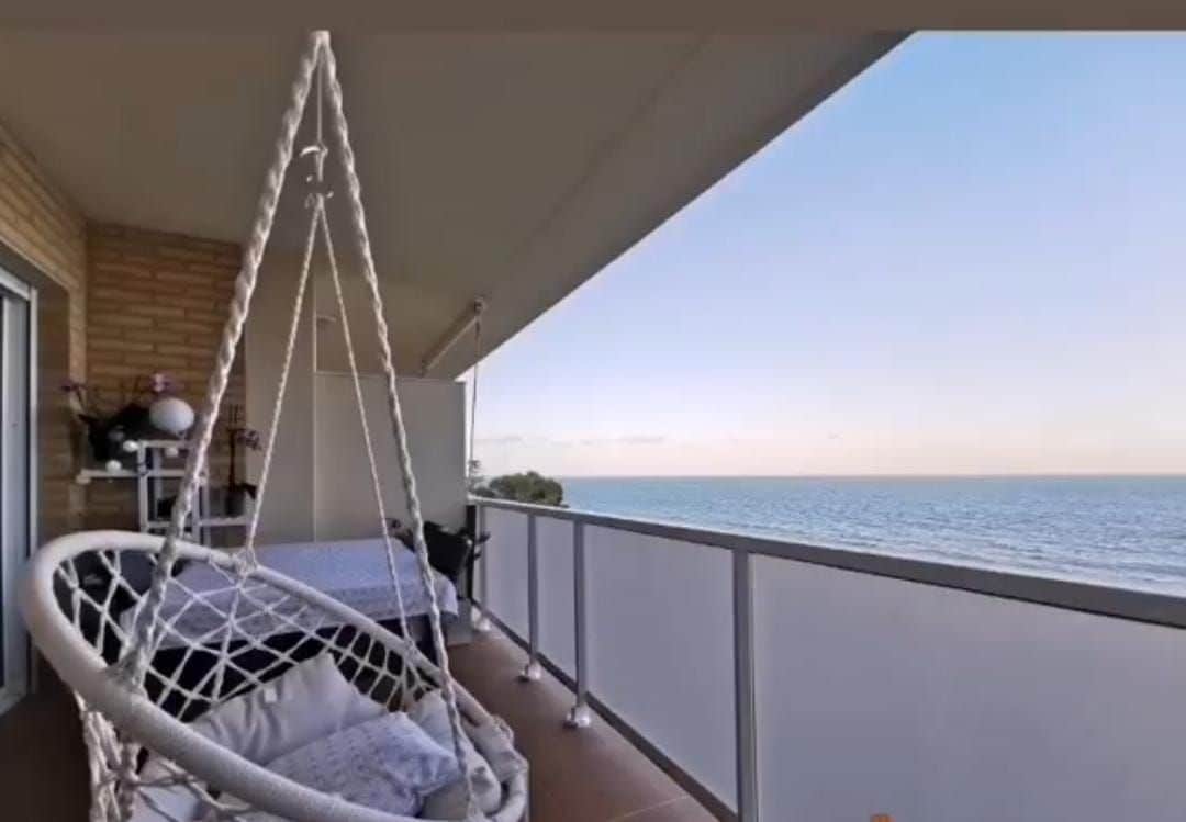 A hanging chair is positioned on a spacious balcony, offering a view of the tranquil sea. The railing frames the horizon, while soft light reflects off the water, enhancing the overall serenity of the outdoor space.