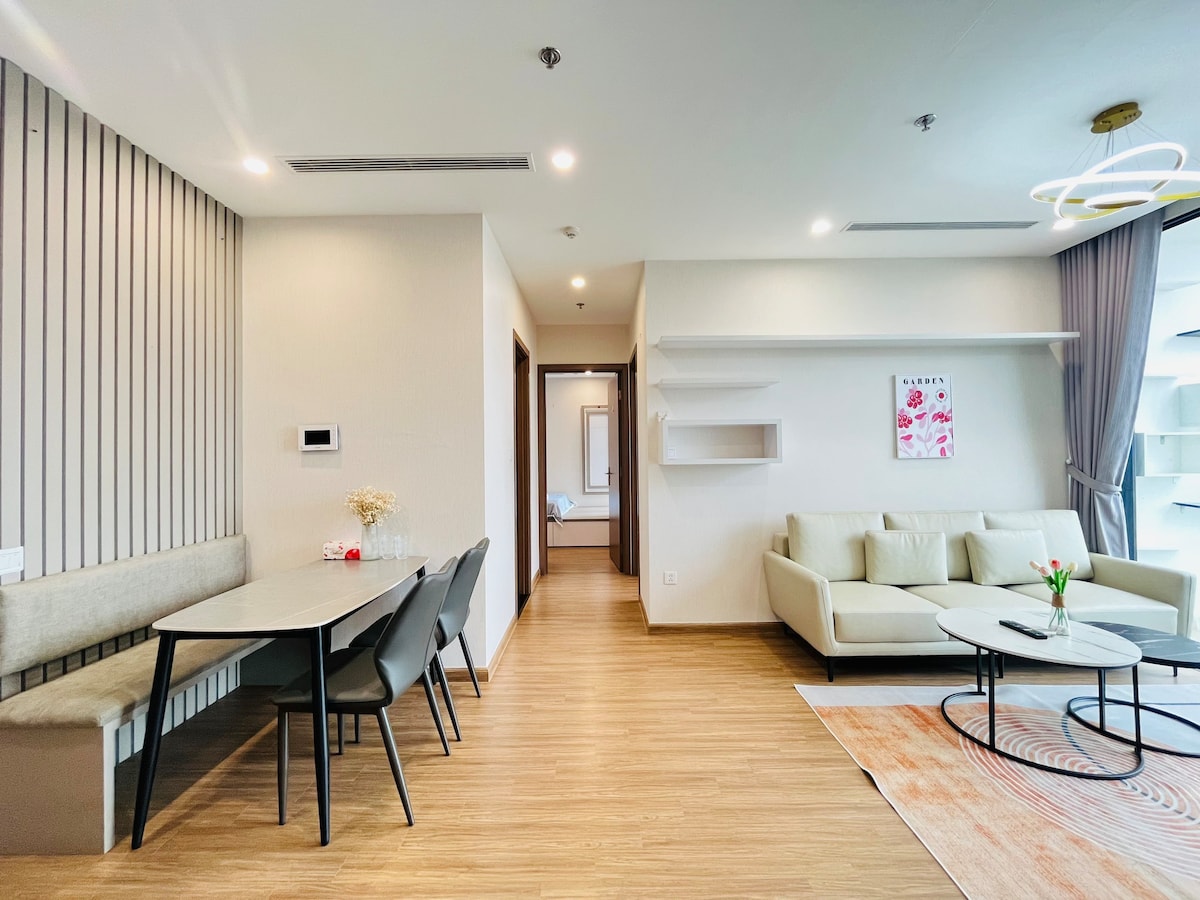 A modern living space features a light-toned sofa and a round coffee table, complemented by a decorative rug. A dining area includes a sleek table with four chairs. Flanked by soft lighting, the walls are styled with wood paneling, enhancing the room's spacious feel.