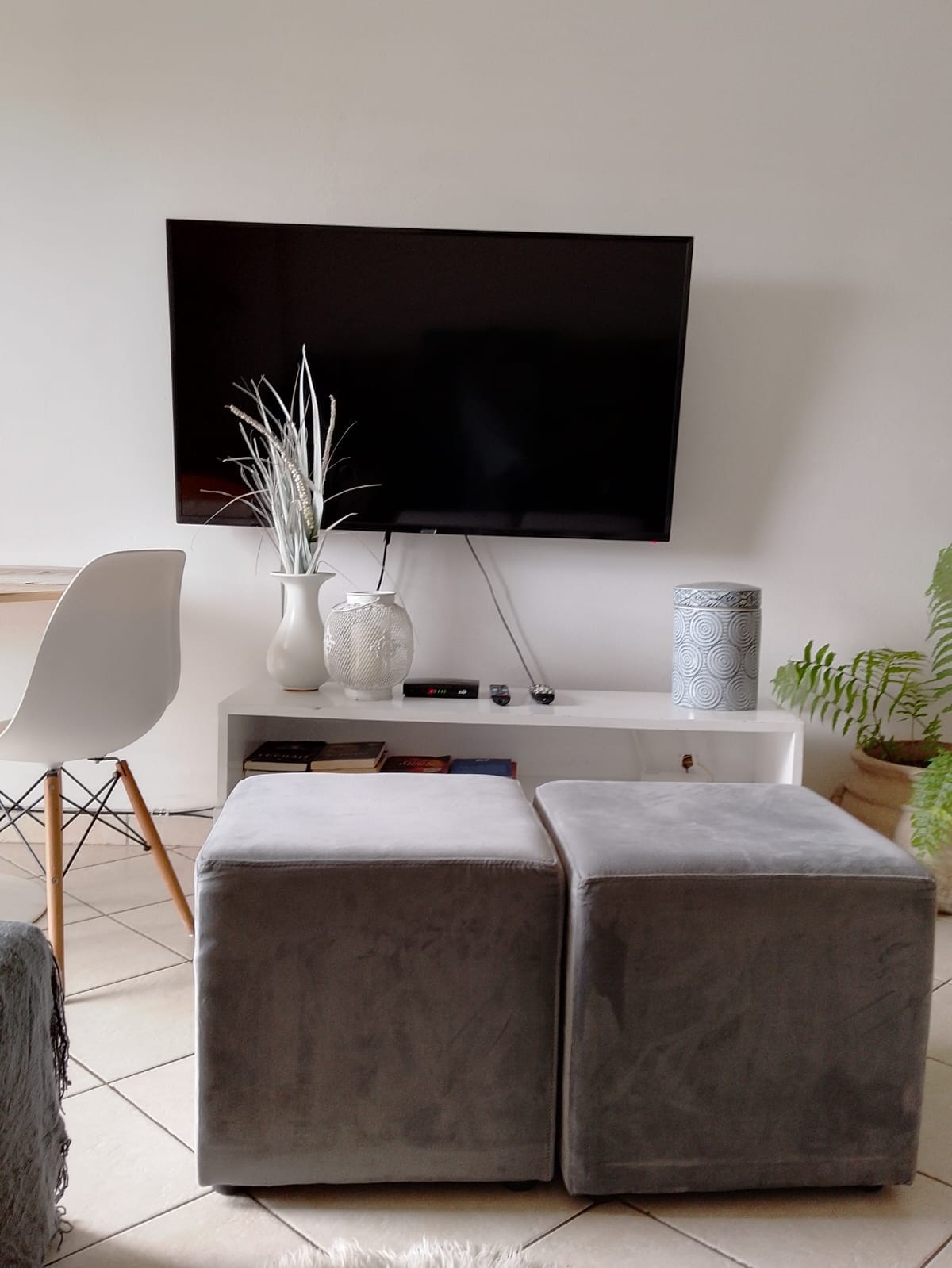 A living area is shown with a flat-screen TV mounted on a white wall. Two gray ottomans are positioned in front of a modern white console table that holds various decorative items. A white vase with dried branches and ferns adds greenery to the space.