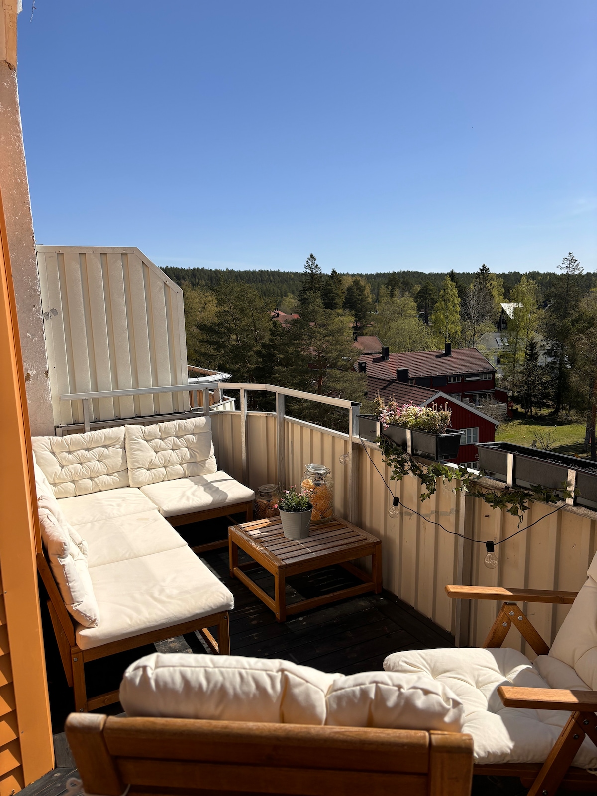 A balcony area features comfortable seating arranged around a wooden table. A potted plant is positioned centrally on the table, with greenery visible in the background. The clear blue sky and distant trees create a serene outdoor space.