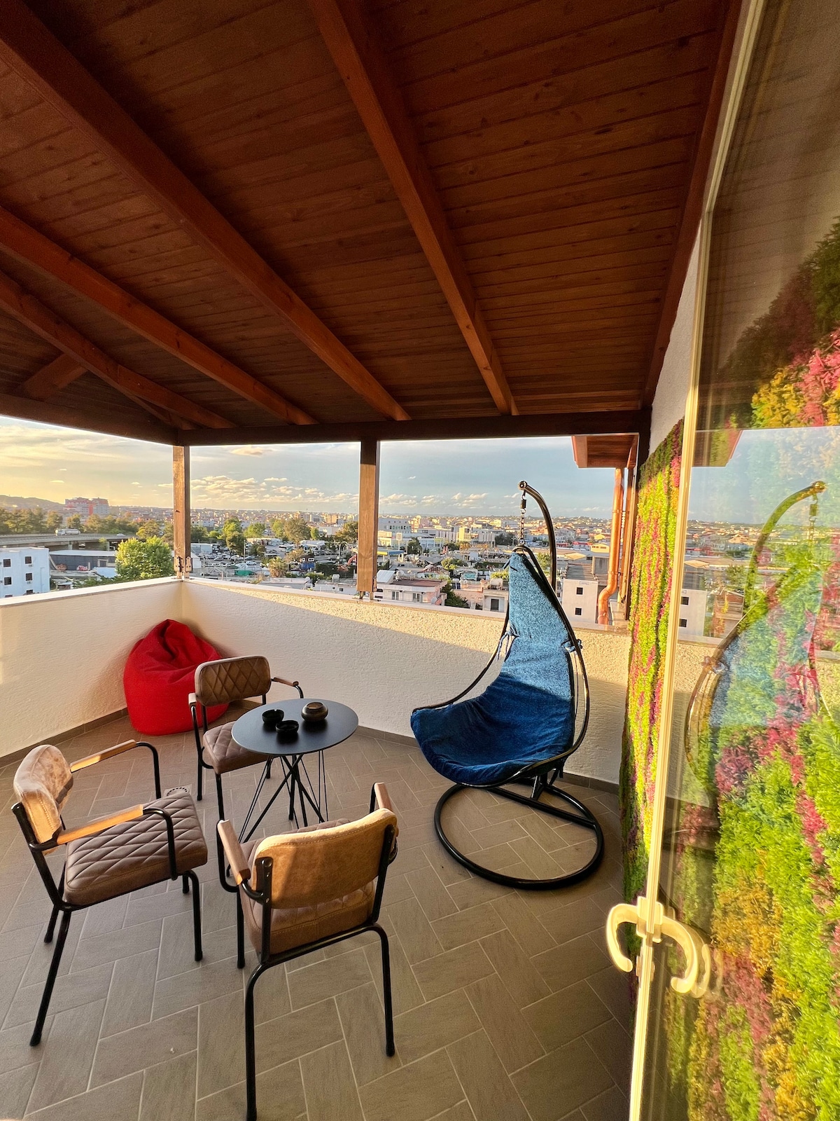 A balcony space is presented with a wooden ceiling and scenic views. A blue hanging chair and four seating options, including two stylish chairs and a bean bag, are arranged around a small table, inviting relaxation. The walls are brightened by vivid green plants.