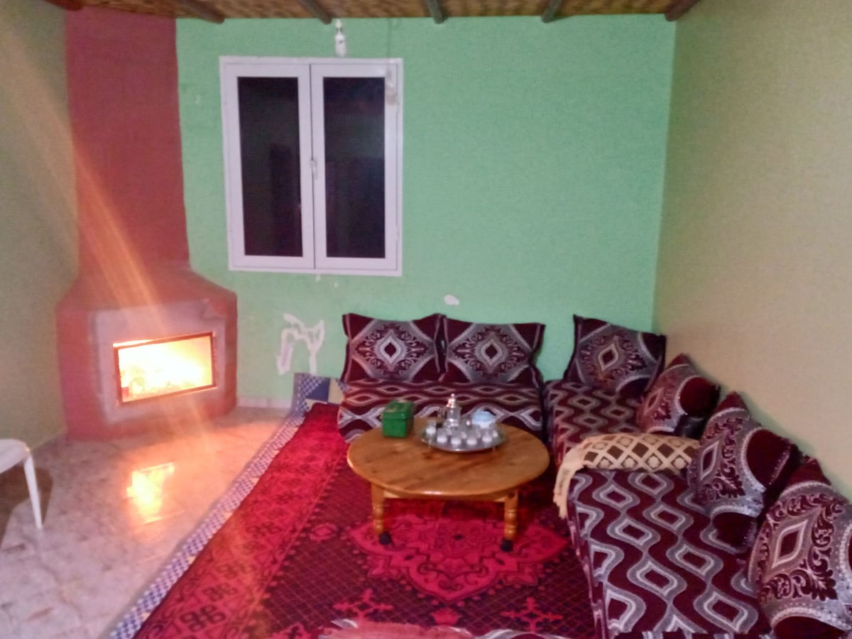 A cozy seating area features a low wooden table surrounded by patterned cushions. A fireplace adds warmth to the space, with a window positioned to allow natural light. The walls are painted in a calming green shade, enhancing the inviting feel of the room.